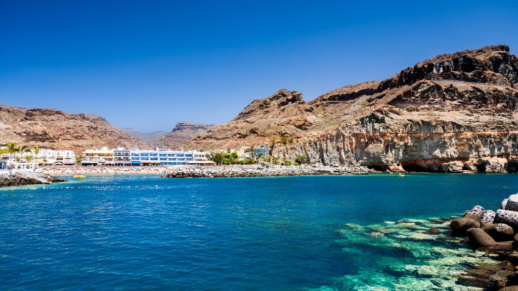 Coastal resort buildings beside a sheltered bay with clear blue sea and rocky cliffs under a cloudless sky