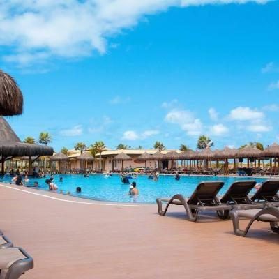 People relaxing in a large outdoor swimming pool with sun loungers and thatched parasols under a bright blue sky