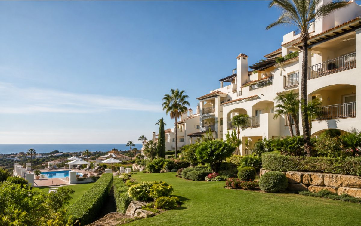 Sunlit hillside resort with white apartment buildings, landscaped gardens and a swimming pool overlooking the sea