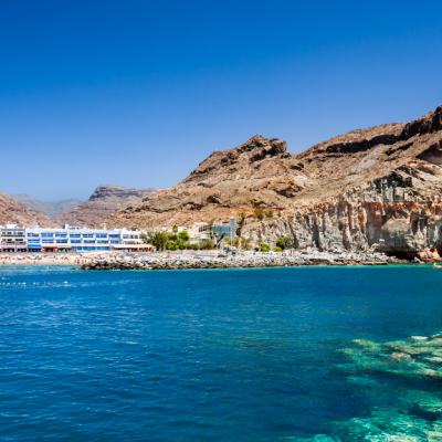 Coastal resort buildings beside a sheltered bay with clear blue sea and rocky cliffs under a cloudless sky