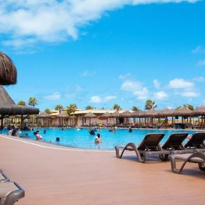 People relaxing in a large outdoor swimming pool with sun loungers and thatched parasols under a bright blue sky